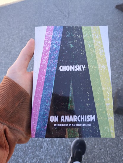 The image depicts a young adult in their 20s standing outdoors.

The person is holding a book titled 'Chomsky On Anarchism'.