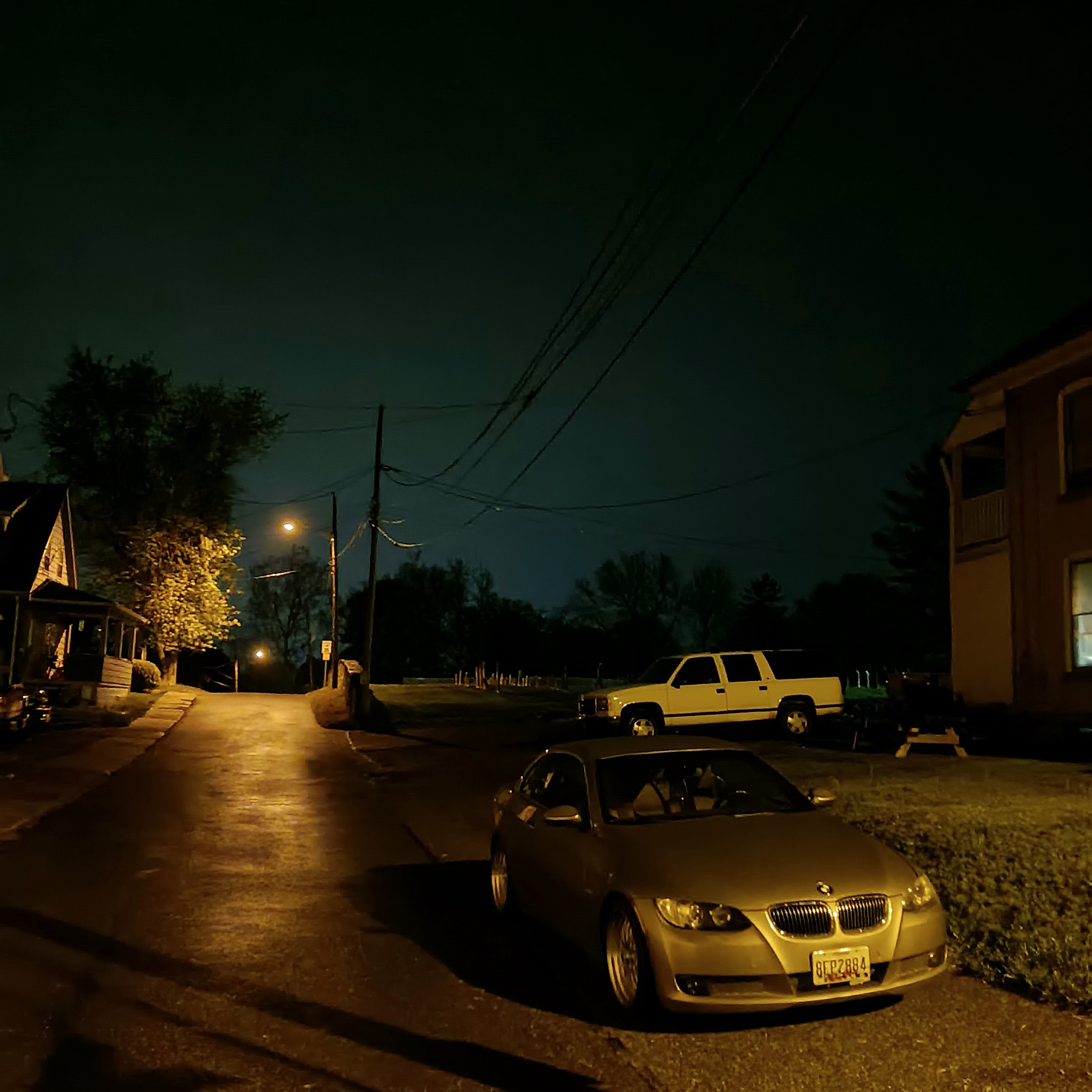 a road up a small lamplit hill at night, cars parked on the side. framed by two houses.