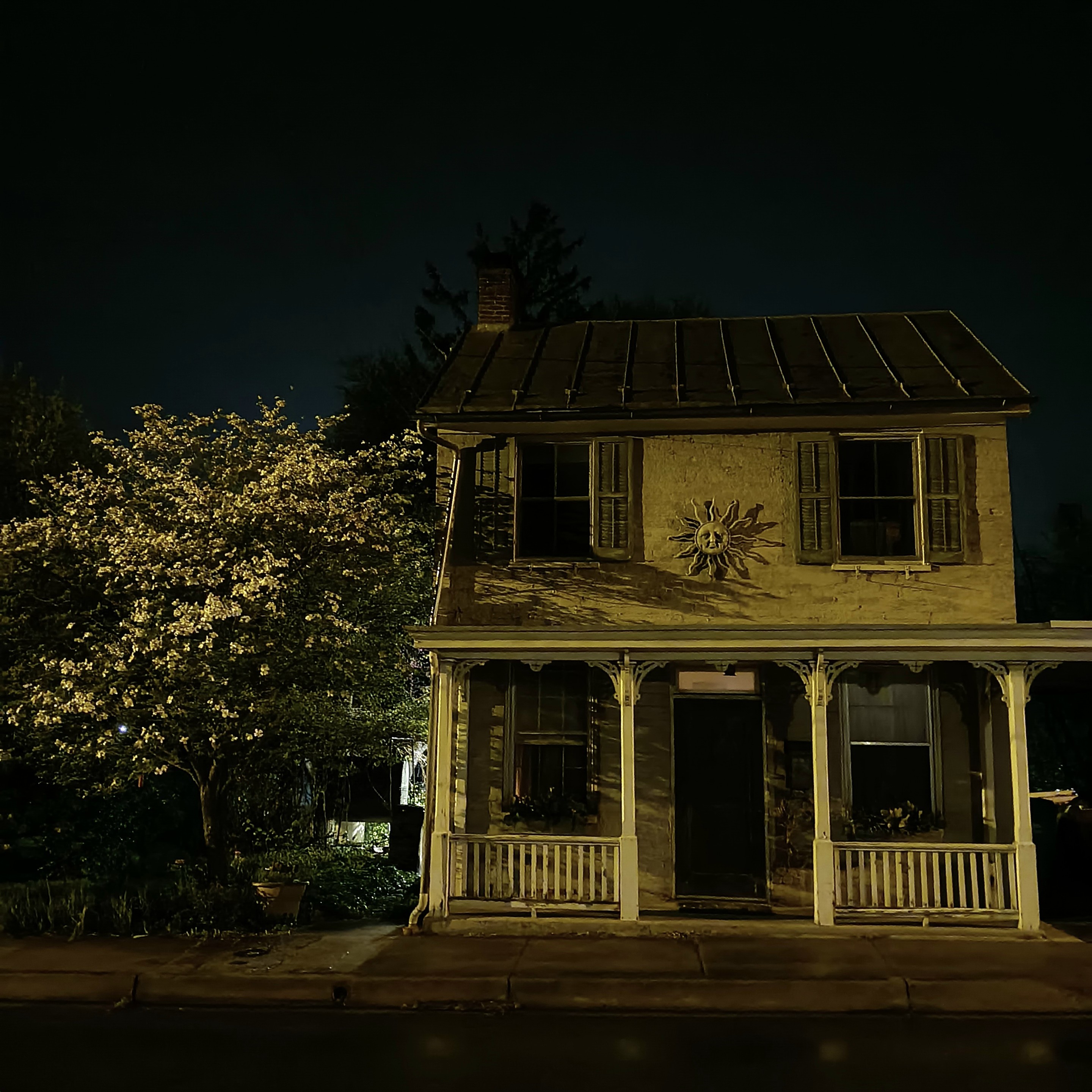 house with a sun decoration on the front, a tree with white flowers grows next to it.