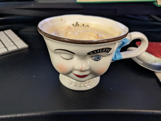 A small ceramic mug shaped like a face. It has features painted on, including red lips, pink cheeks, and a winking eye. The open eye is blue and has an extra thick eyebrow that says "BAILEYS'". In the cup is ice cream and espresso. Painted in the inside of the cup is the word "yum."