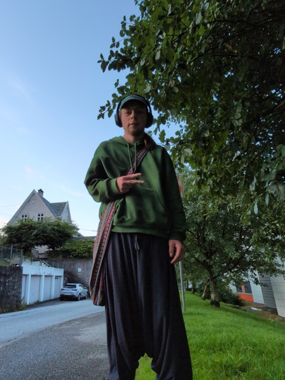 A person in a green hoodie and loose dark pants stands on a sidewalk near trees and houses, making a peace sign with their right hand.