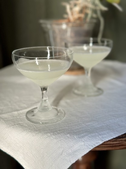 Two elegant coupe glasses filled with a light-colored drink sit on a textured white tablecloth. A blurred plant is visible in the background.