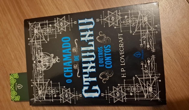 imagem mostra a capa de um livro intitulado "O Chamado de Cthulhu e Outros Contos" de H.P. Lovecraft. A capa é predominantemente preta, com detalhes em branco e azul. O título "O Chamado de Cthulhu" está em letras grandes e estilizadas, com um design que lembra hieróglifos ou símbolos místicos. Ao redor do título, há desenhos geométricos e símbolos que parecem ser de origem ocultista ou esotérica, incluindo estrelas e círculos. O nome do autor, H.P. Lovecraft, está escrito em letras menores, na…