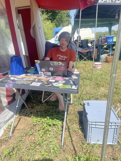 Ein Typ sitzt auf einem Festival Campingplatz vor einem Zelt und schaut auf seinen Laptop. Am Tisch steht eine Kaffeekanne und ein Drumstick.