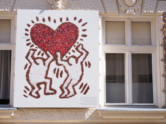 Zwischen zwei Fenstern an einem Haus ein Foto. Im Stil von Kaith Haring zwei vergnügte Personen unter einem roten Herz. Wenn man das Foto genau betrachtet erkennt man die Zusammensetzung, das rote Herz wird aus Hagebutten gebildet, die Striche aus Erde und der Hintergrund ist Schnee. Das Bild ist Teil der Ausstellung La Gacilly Baden - Pia Moana Scharler & Craig Dillon "Der Wald ist der größte Künstler"