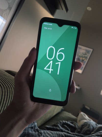 A hand holds a smartphone displaying the time 06:41 with a green background. The phone is in airplane mode. The setting appears calm, with a couch and artwork in the background.