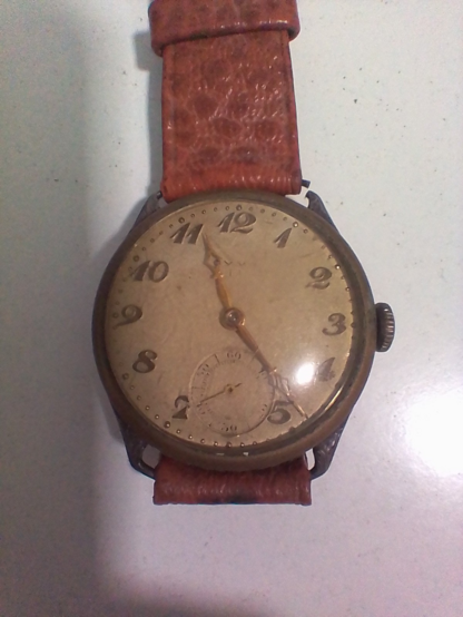 Se ve un reloj de esfera y letras doradas. Tiene el segundero aparte de minutero y horas. No se ve la marca. Tiene una correa de lo que parece ser cuero marrón. La sujección de la correa está rota en uno de los extremos.