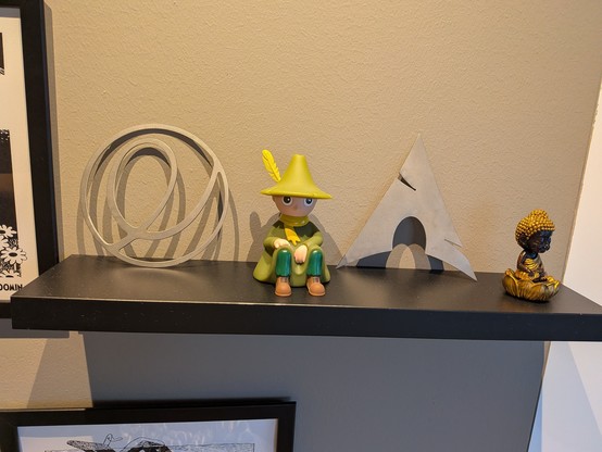 A black shelf displays a figurine in a green cloak and yellow hat, flanked by circular and triangular metal sculptures, alongside a small meditating Buddha statue.