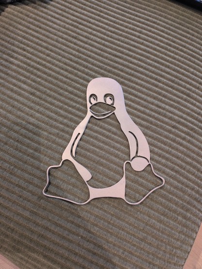 A metal cutout of a penguin lies on a textured, ribbed gray surface. The penguin is stylized and adds a playful tone to the minimalist setting.