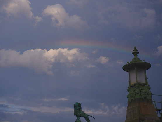 sky with storm clouds and a rainbow, on the edge a lion sculpture in the center  and a lamp on the right