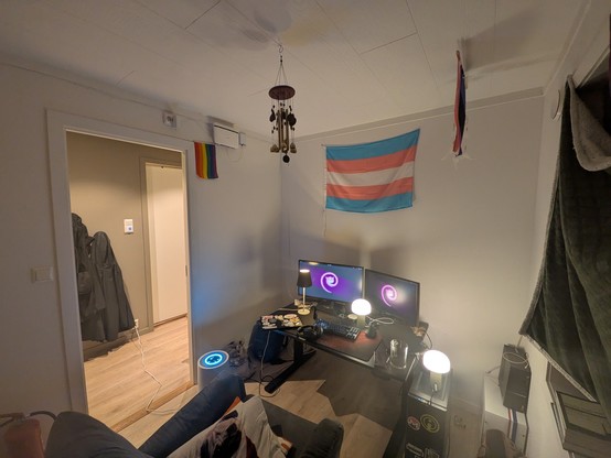 A cozy room with a computer setup on a desk; two monitors with glowing neon Debian swirl, surrounded by small lamps. A transgender pride flag and a small rainbow flag hang on the wall, creating a supportive and positive atmosphere.