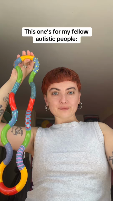 Person holds a colorful, twistable sensory toy with a joyful expression. Caption reads, "This one's for my fellow autistic people." Tattoos visible on arm.