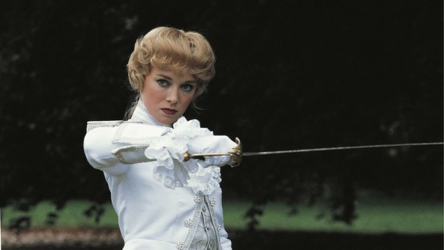 A woman in a white 18th-century outfit holds a fencing stance with a focused expression. The background is dark and blurred, enhancing her determined pose.