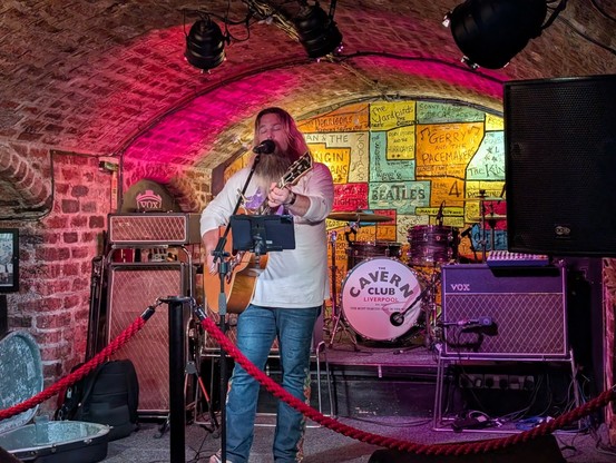 A man, with more than a passing resemblance to Jack Black, performs on a guitar in front of the iconic wall at The Cavern Club 

The lighting and wall decoration combine to give a rainbow effect, fitting for Pride.