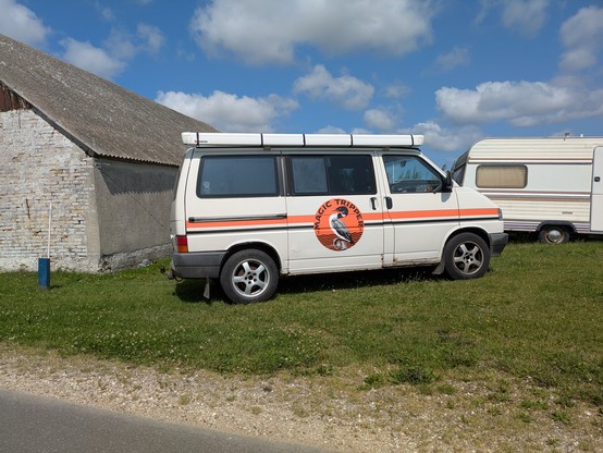 Foto eines Campervans von der Seite, auf einer Wiese neben der Strasse stehend. Links davon eine Scheunenwand, rechts im Hintergrund ein Wohnwagen. Darüber blauer Himmel mit lebhaften, weißen Wolken.
Auf dem Campervan prangt ein Aufkleber mit der Aufschrift 'Magic Tripper'.