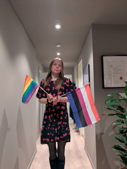 Young adult standing in a hallway holding a rainbow flag and a genderfluid flag, wearing a floral dress with a neutral expression; background features simple decor and a potted plant.