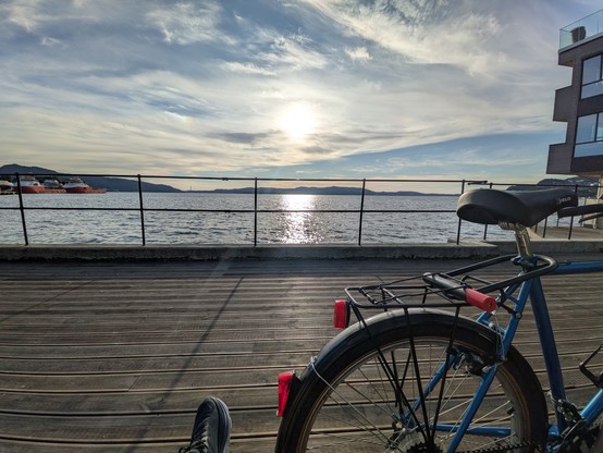 A bicycle rests on a wooden pier at sunset, overlooking a calm sea. The sky is partly cloudy, and distant boats are visible. The scene is peaceful and serene.