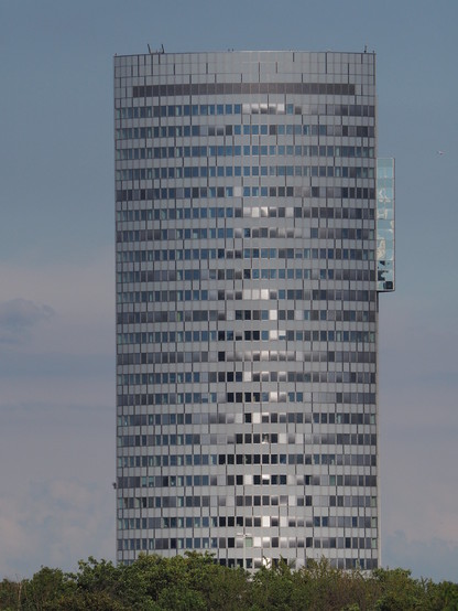 Ein Hochhaus-Turm (etwa 25 Stockwerke) isoliert in der Landschaft, am unteren Rand grüne Laubbäume.
