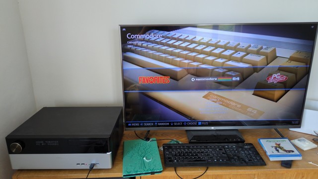 Flaches Computergehäuse neben Fernseher. Darauf zu sehen ist der Batocera startbildschirm auf dem gerade Commodore 64 ausgewählt ist