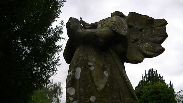 a headless and damaged stone angel.  dull clouds in the background. and some trees.