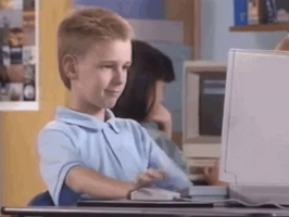 A captivating image displays a young boy in a striking blue shirt, intently gazing at a computer screen. His vibrant red hair stands out against the desks clutter, where a keyboard, mouse, and monitor reside. The background blurs into a cozy room with a yellow wall and blue door, capturing the essence of a tech-savvy teenagers focus.