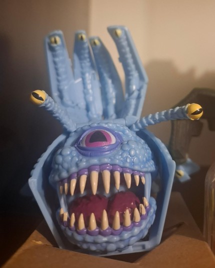 Fotografia de um bonequinho de plástico de um Beholder, um monstro do dungeons and dragons cujo formato de corpo é essencialmente uma bola com uma grande boca, que está aberta e cheia de dentes afiados, e um grande olho. Além disso vemos uns tentáculos nas pontas dos quais há olhos menores.