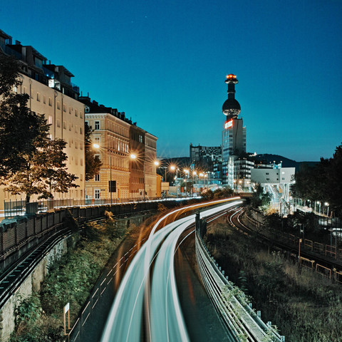 Nachtaufnahme in der Stadt. Man sieht die oberirdisch verlaufende U-Bahn als Lichtspuren und einige Straßenlaternen mit Blendensternen, im Hintergrund die Müllverbrennungsanlage Spittelau, welche von Hundertwasser entworfen wurde.
