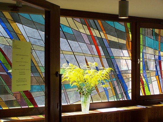 Eine horizontale Fensterreihe aus dem Innenraum fotografiert. Bunte Bleiglasfenster, in der Mitte eine Blumenvase mit gelben Blüten.