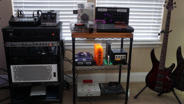 A picture showing (from left-to-right): a 19" audio rack with a mixer and microphone amp on top, a compresser/expander (dbx 286s), a shelf holding miniDiscs and a mini PC, a TASCAM MD-350 MiniDisc player, a 4U rack-mounted PC (my main rig), and a Sony MDS-E12 MiniDisc player.  A shelf full of retro consoles including an NES, Sega Master System, PS1, GBA (top shelf), SNES, Dreamcast, PS2, and Gamecube (middle shelf), Sega Saturn, Sega Genesis/Mega Drive (bottom shelf).  Beside the shelves are tw…