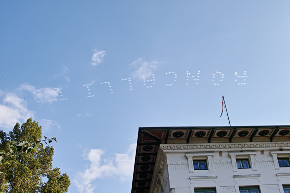 Blick in den blauen Himmel, quer über das Bild die Buchstaben RONCALLI. Am unteren Bildrand noch ein Gebäude und ein Baum.