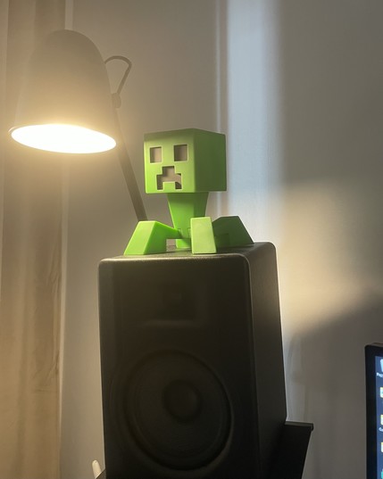 A green Minecraft Creeper figurine sits on top of a black speaker, beside a lamp that casts warm light.
