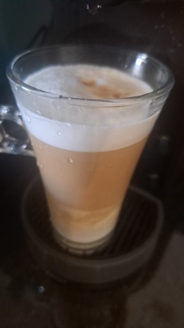 A imagem mostra uma xícara de vidro transparente mostrando um Cappuccino em seu interior. A xícara está posicionada sobre um suporte de máquina de café, com uma camada de espuma branca no topo, seguida por uma camada mais clara de café e uma camada mais escura na base. A xícara tem uma alça transparente e está parcialmente coberta por gotas de condensação, indicando que a bebida é quente. O fundo é escuro, com uma máquina de café visível, sugerindo que a bebida foi preparada recentemente.
A apr…