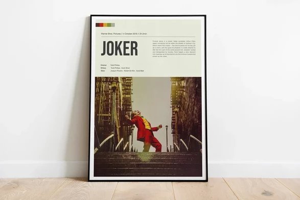 Framed "Joker" poster on floor, showing a clown-costumed figure dancing on stairs in a city. Bold text and muted tones create a dramatic feel.