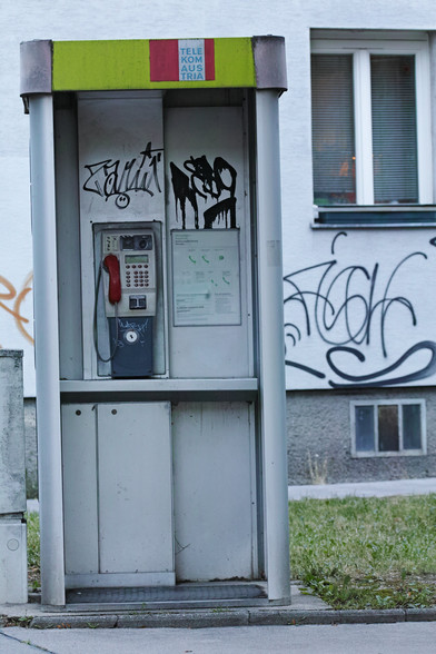 Eine für das Jahr 2000 typische österreichische graue Telefonzelle, die Tür vorne fehlt. Der Telefon-Hörer ist rot, oberhalb vom Telefon sind Graffiti-Schriften und auch auf dem Gebäude dahinter.