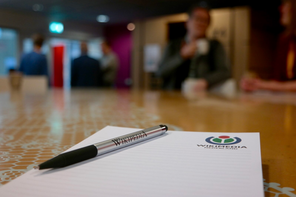 A metallic pen with “Wikipedia” printed on it rests on a notepad bearing the Wikimedia Nederland logo, placed on a patterned table. In the blurred background, several people stand and converse in a conference-like setting.