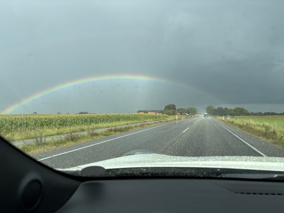 A strong rainbow from a car cabin against a dark grey sky. The window is lightly covered by raindrops. Green fields are on either side of the road. 
