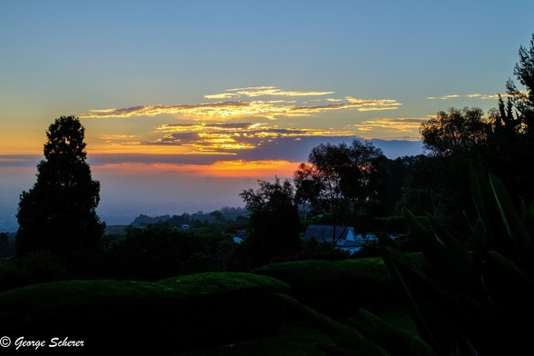 Clouds in the distance against an otherwise blue sky are illuminated from behind in orange by the sun.  in the foreground are the dark silhouettes of trees.