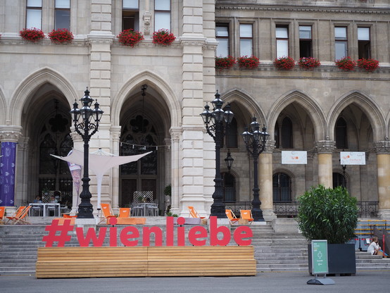 Die Gebäudefront mit Spitzbögen unten und eine Fensterreihe, vor den Fenstern Blumenkästen mit roten Geranien.  Vor dem Gebäude drei schwarze Lampen. Im Vordergrund eine Sitzbank aus Holz, die Lehne der Bank trägt den Schriftzug #wienliebe. 