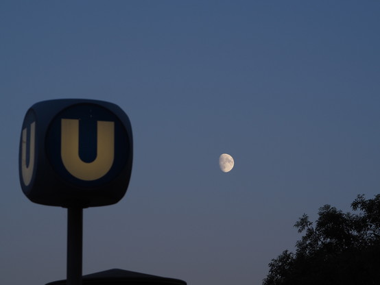 Links ein blauer Würfel Hinweisschild für die U-Bahn, rechts ziemlich klein, als hellster Punkt im Bild,  der Mond und ein Baum am unteren Bildrand. Der Himmel ist wolkenlos.
