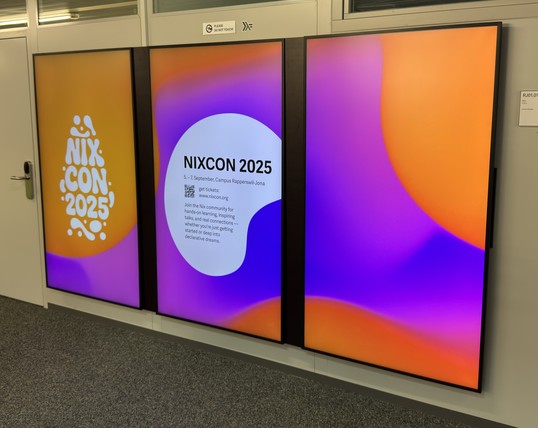 Photo of digital signage monitors displaying the NixCon 2025 logo and dates. 