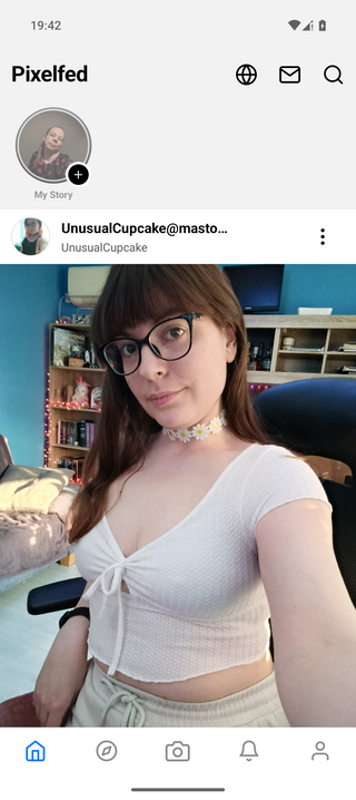 A person with glasses and long hair takes a selfie in a cozy room with bookshelves and a blue wall. They wear a white top and floral choker.