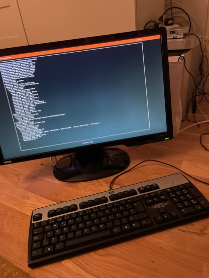 A computer monitor displays command line interface text, while a keyboard sits in front of it on a wooden surface. The background includes a glimpse of a room with some cables and a white object.