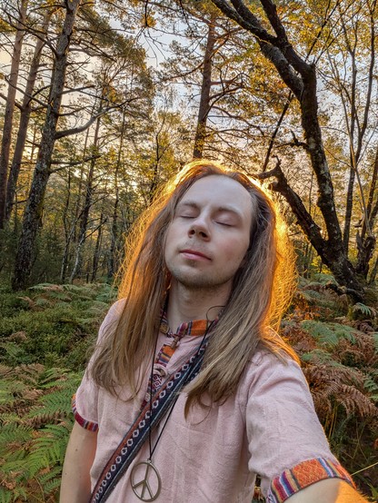 A person with long hair, wearing a pink shirt and peace symbol necklace, stands in a sunlit forest with eyes closed, appearing serene and content.