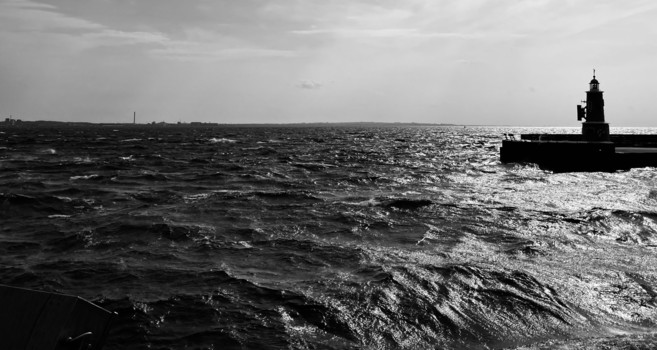 A black and white image of a turbulent sea with visible waves under a cloudy sky. A lighthouse sits on a pier in the foreground, with a distant shoreline visible in the background. The scene conveys a dramatic and moody atmosphere.