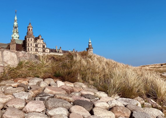 A historic castle with various towers is visible against a clear blue sky, surrounded by grassy hills and a rocky foreground.