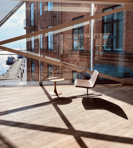 A modern interior space featuring a stylish chair and a small round table. There are large glass walls showcasing a view of a dock with boats, while sunlight creates dynamic shadows on the wooden floor. The backdrop includes a brick building, highlighting a blend of contemporary