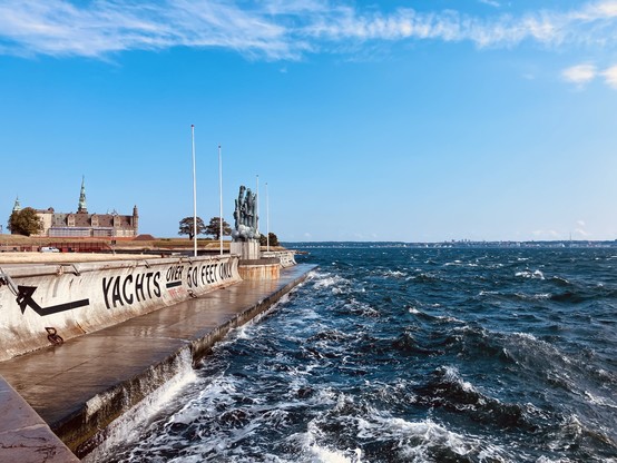 A harbor scene featuring a wave-washed concrete pier with a warning sign about yacht sizes. In the background is a historic castle, while the ocean stretches into the distance under a blue sky with scattered clouds.