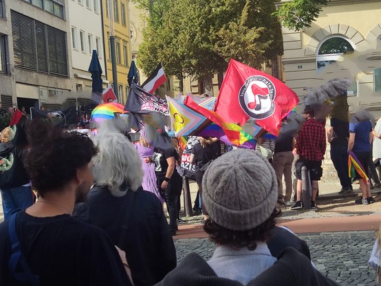 Auf dem Foto ist eine Straßenszene bei einer Demonstration oder einem CSD-Umzug zu sehen. Viele Menschen stehen dicht beieinander auf dem Gehweg. Einige tragen bunte Kleidung, manche auch schwarze Outfits.

Mehrere Fahnen sind gut erkennbar:

Im Vordergrund eine große rote Fahne mit einem Kreislogo und der Aufschrift „Love Music – Hate Fascism“.

Daneben eine Progress-Pride-Flagge (Regenbogen mit zusätzlichen Farben für Trans* und PoC).

Weiter hinten eine klassische Regenbogenflagge sowie eine…