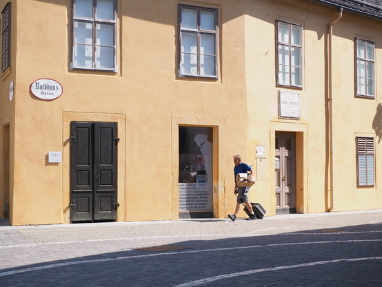 In einer Straßenszene sieht man eine Person mit Rollkoffer und einem Bündel Papier unter dem Arm an einem Gebäude vorbeighen. Ein Schild an der Wand mit der Aufschrift "Rathaus-Gasse".