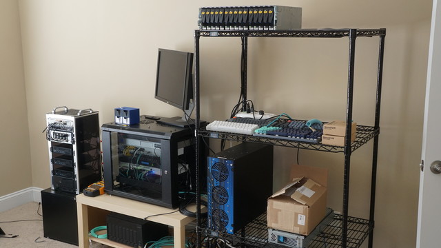 A picture of the homelab. From right-to-left: the minirack hosting all of the compute, a small network rack above a backup NAS, some black shelving holding the primary NAS along with the EMC shelf and some keyboards.